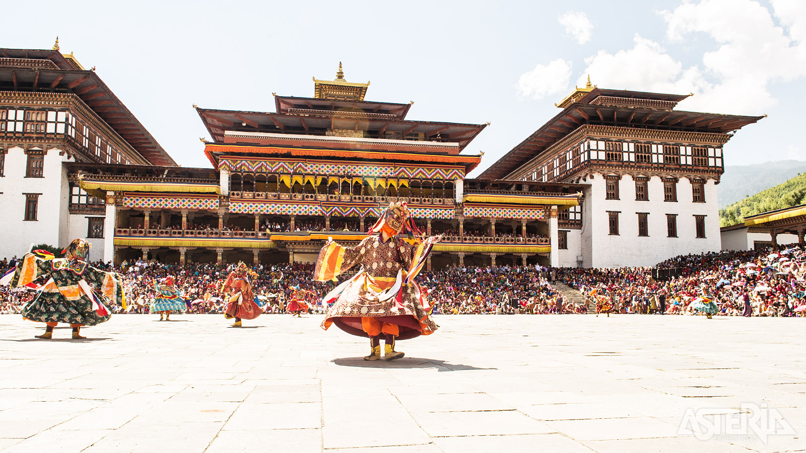 Gemaskerde dansers met zijden kostuums dansen en springen tegen een achtergrond van hemel en bergen tijdens het Thimphu Festival