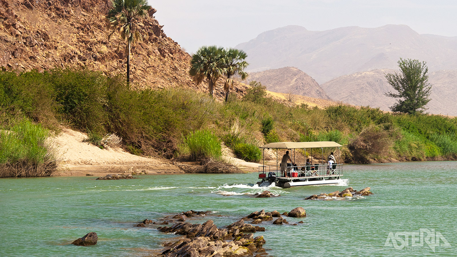 De logies van het Okahirongo River Camp kijken uit over de Kunene-rivier, het prachtige gebergte en wuivende palmbomen